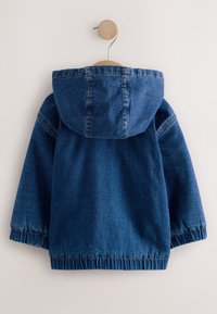 Giacca con cappuccio in denim blu scuro con polsini e orlo elastici appesa a un gancio rotondo in legno sulla parete.