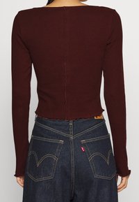 Top de manga larga de color rojo oscuro con cuello recto y dobladillo desigual. Presenta una costura central visible y bordes con forma de concha.