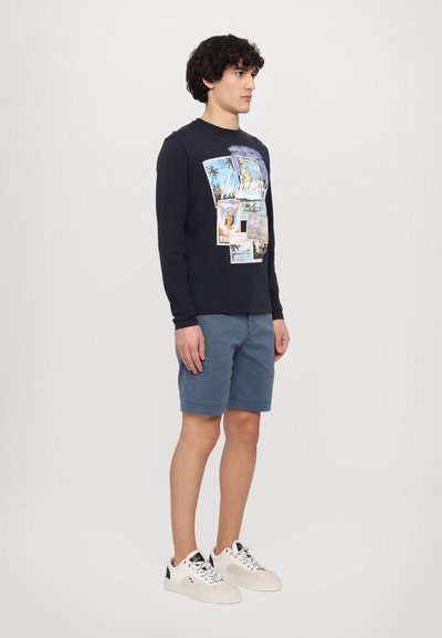 Camisa de manga larga azul marino con un estampado gráfico que presenta tarjetas postales; combinada con pantalones cortos de color azul claro. El modelo lleva zapatillas blancas y negras.