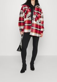 Mujer con chaqueta de cuadros oversized en rojo, blanco y marrón, top negro con cremallera, jeans skinny negros, botas de tacón negras, sosteniendo un pequeño bolso negro.