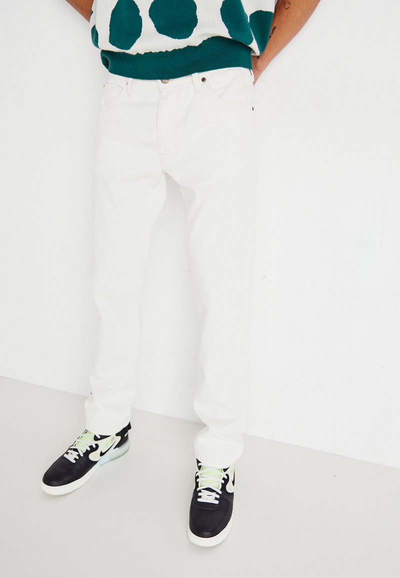 White denim jeans with a slim fit, paired with black sneakers featuring green accents. Background is plain white for contrast.