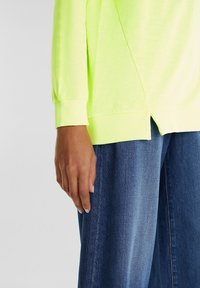 Top de manga larga verde neón con puños acanalados, combinado con unos vaqueros de denim azul claro de corte recto y textura sutil, de ajuste holgado.