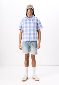 Short-sleeve plaid shirt in light blue and white, paired with light denim shorts. Worn with a beige cap and white socks, featuring casual sneakers.