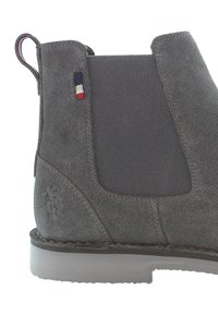 Botín Chelsea de ante gris con paneles laterales elásticos, superficie texturizada y un logo sutil en el talón. Presenta acentos en rojo, blanco y azul.