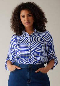 Camicia azzurra e bianca con motivo a righe, con maniche arrotolate e colletto. Abbinata a una gonna di jeans, con le mani nelle tasche.