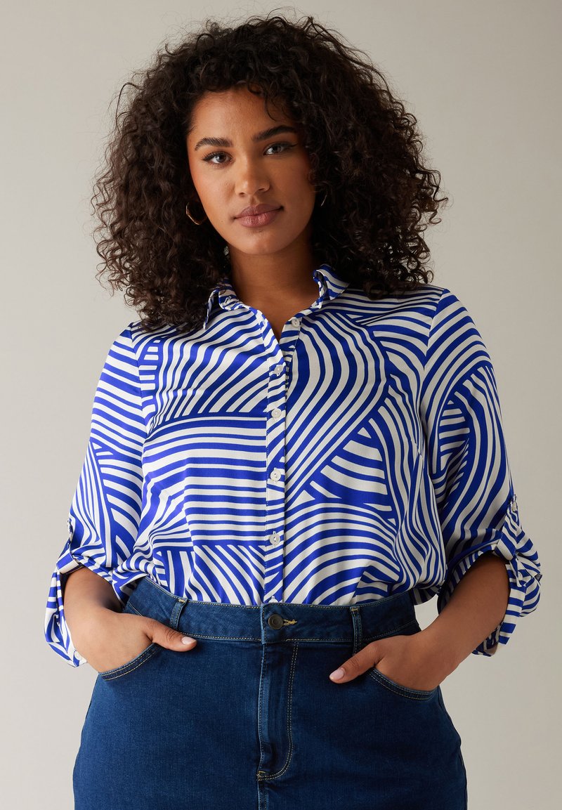Camicia azzurra e bianca con motivo a righe, con maniche arrotolate e colletto. Abbinata a una gonna di jeans, con le mani nelle tasche.