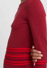 Personne portant un haut à manches longues rouges avec des rayures horizontales rouges et rouge foncé au niveau de la taille, se tenant le côté d'une main contre un fond uni.