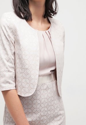 Femme portant une veste rose clair à motifs à manches trois-quarts par-dessus une robe assortie avec un col plissé.