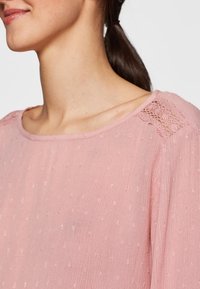 Hellrosa Bluse mit strukturiertem Stoff und winzigen Punkten, einem weiten Ausschnitt und filigranen Spitzenakzenten an einer Schulter.