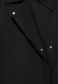 Prim-plan al reverului unei jachete negre cu trei capse argintii vizibile și cusături detaliate pe material.