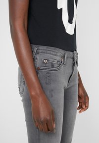 Szare dżinsy z przetartymi detalami, mosiężnymi nitami i dopasowanym krojem. W komplecie czarna koszula z białym wzorem graficznym.