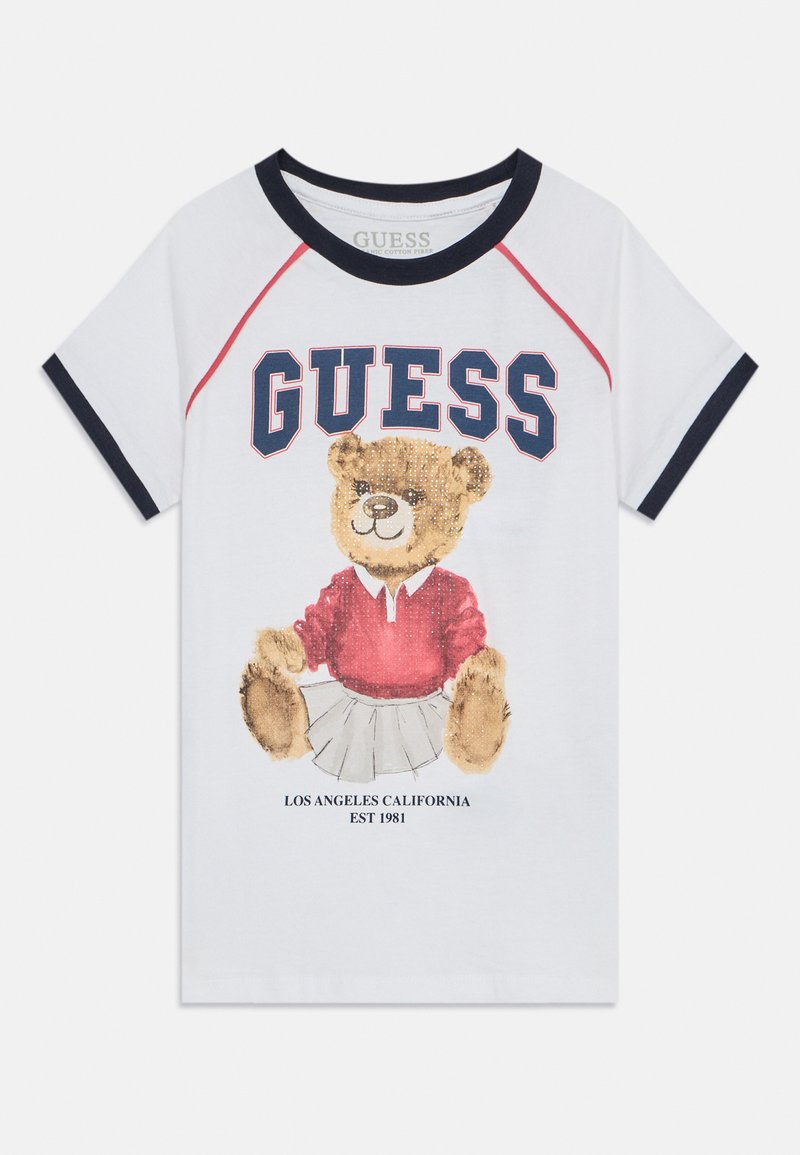 Camiseta de algodón con base blanca, ribete azul marino y acentos rojos. Presenta un gráfico de un oso de peluche con una camiseta y falda rojas, con el texto "GUESS" arriba.