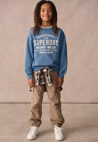 Felpa blu con testo bianco, pantaloni cargo beige e sneakers bianchi. Camicia a quadri annodata in vita. Look casual e rilassato.