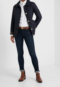 Marinblå vadderad jacka med gyllene knappar, bärs över en vit skjorta, tillsammans med mörka jeans och bruna läderankelstövlar.