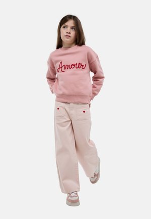 Fille en sweat-shirt rose avec le texte "Amour", pantalon large rose clair avec des détails en forme de cœur, et baskets rose et blanc, marchant les mains dans les poches.