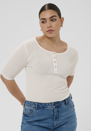 Vrouw met donker haar, gekleed in een wit topje met knopen en blauwe spijkerbroek, kijkt zijwaarts met haar armen achter haar rug gestoken.