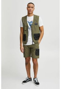 Vesta utilitară verde măsliniu cu două buzunare mari în față; asortată cu un tricou alb cu un design grafic albastru și pantaloni scurți verzi măslinii.