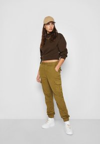 Hnedý skrátený sweatshirt, olivové cargo nohavice s bočnými vreckami a biele tenisky. Oblečenie doplnené béžovou čiapkou.