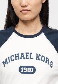 T-shirt branca com mangas raglan azul marinho, apresentando "MICHAEL KORS" e "1981" impressos em texto azul marinho negrito na frente.