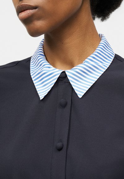 Približen posnetek osebe, ki nosi mornarsko modro srajco z gumbi iz enakega blaga ter modro-belim črtastim ovratnikom z ostrim robom.