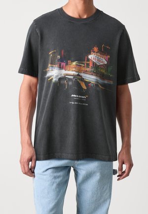 Personne portant un T-shirt noir McLaren Formula 1 avec un graphisme de voiture de course et un panneau au néon de Las Vegas, associé à un jean bleu clair.