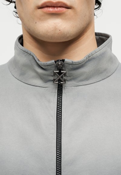 Κοντινό πλάνο του κάτω μέρους προσώπου και του λαιμού ενός ατόμου που φοράει ανοιχτογκρι ζακέτα με φερμουάρ σε μαύρο και μεταλλικό τανταλάκι.