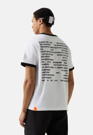 Mand iført hvid T-shirt med sort tekstblok og sort kant på ærmer og krave, kombineret med sorte bukser og sort kasket på hovedet.