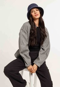 Giacca oversize grigia in tessuto morbido, abbinata a un top nero e pantaloni neri larghi. Un cappello a secchiello in feltro blu completa il look.