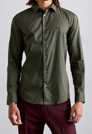 Uomo che indossa una camicia verde oliva aderente con bottoni e maniche lunghe, abbinata a pantaloni rosso scuro, su uno sfondo semplice.