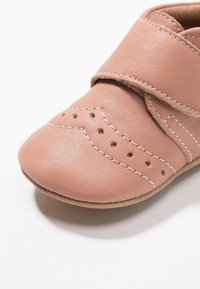 Zapato de cuero rosa claro con puntera redondeada, correa de velcro, perforaciones decorativas y costuras en blanco. Suela de ante suave.