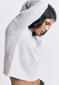 Žena si upravuje svetlosivý sveter, pričom odhaľuje časť svojho holého brucha a má dlhé tmavé vlasy na jednoduchom bielom pozadí.