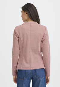Blazer de punto rosa con espalda entallada, cuello con muesca y tejido texturizado. Combinado con jeans de mezclilla azul, mostrando detalles de costura.
