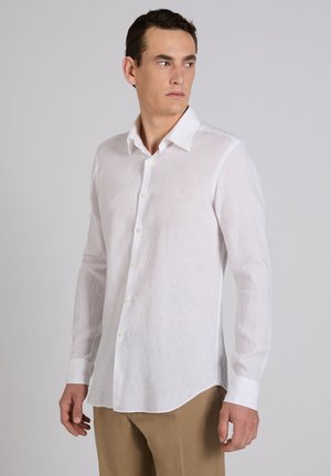 Mężczyzna w białej, długorękawowej koszuli zapinanej na guziki i beżowych spodniach, patrzący w lewo na jednolitym, szarym tle.