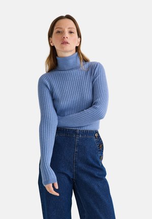 Pull à col roulé côtelé bleu à manches longues, associé à un pantalon en jean taille haute avec boutons sur le devant. Texture lisse, silhouette ajustée.