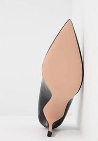 Chaussure à talon haut noire en cuir à bout pointu, positionnée avec la semelle vers le haut contre un mur blanc d’angle, montrant une semelle extérieure beige lisse et un talon aiguille.