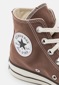 Zapatilla alta de lona marrón con ojales blancos, costuras en contraste, suela de goma y un parche de logo circular en el lado.