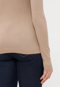 Beige long-sleeve top with ribbed cuffs and hem, made of soft knit fabric. Worn with dark blue fitted jeans. No visible patterns or hardware.