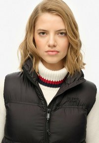 Junge Frau mit blonden Haaren, die einen weißen Pullover mit rot und schwarz gestreiften Mustern sowie eine schwarze, enganliegende Steppweste trägt.