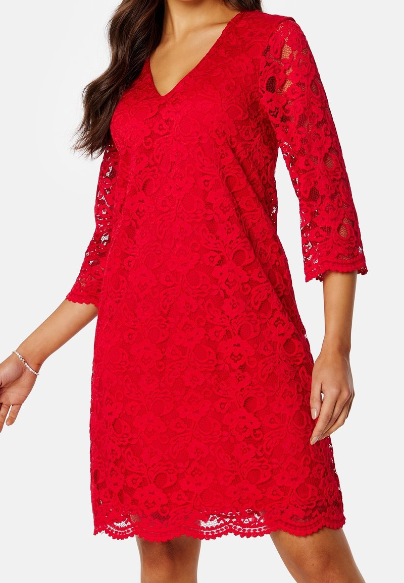 Femme portant une robe en dentelle rouge avec des manches trois-quarts et un ourlet festonné, montrant un motif floral en dentelle et un décolleté en V.
