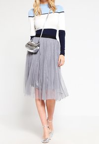 Žena v modro-bílé svetrkové kombinaci, šedé plisované midi sukni, stříbrné crossbody kabelce a stříbrných sandálech na kotníkovém pásku.
