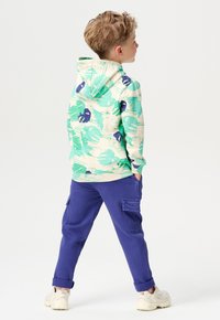 Hoodie met groen, blauw en beige bladpatroon; gecombineerd met paarse cargo broek; casual ontwerp, katoen materiaal, ontspannen pasvorm.