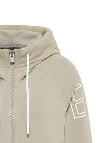 Felpa con zip in tessuto beige chiaro con una trama liscia. Presenta un colletto alto, lacci bianchi e un logo stampato sul maniche.