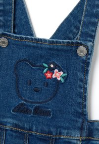 Le salopette in denim presentano un volto d'orso ricamato in blu scuro, adornato con accenti floreali colorati. Completate con hardware argentato e cuciture a contrasto.
