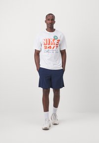 Valge puuvillane T-särk, millel on julge oranž ja roheline Nike bränding, koos tumesiniste lühikesed pükste, valgete sokkide ja heledate sportlike kingadega.