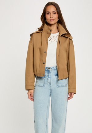 Vrouw draagt een beige jasje met knopen over een wit shirt met uitsnijdingen en lichtblauwe, hooggetailleerde spijkerbroek, staand tegen een effen achtergrond.