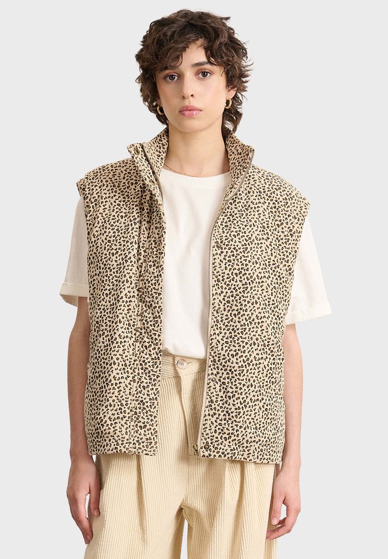 Beige ärmlös väst med leopardmönster, dragkedja fram och hög krage. Bärs över en vit t-shirt; ihop med ljusfärgade plisserade byxor.