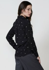 Žena s dugom smeškom smeškom gustom smeškom smeškom brinete s bočne strane, nosi crnu majicu s srebrnim uzorcima zvezda, u kombinaciji sa sivim pantalonama.