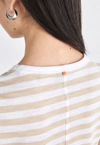 T-shirt rayé avec des rayures horizontales beiges et blanches, doté d'un col rond et d'un petit accent orange à l'arrière du col.