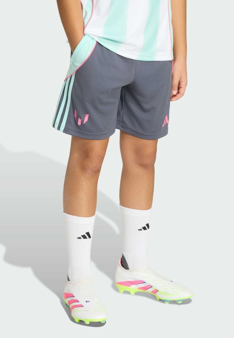 Dítě oblečené do šedých sportovních kraťasů s tyrkysovými pruhy, bílých fotbalových kopaček s růžovými detaily, bílých ponožek a světle modrého a bílého dresu.