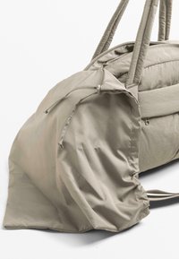 Beige kangaskassi, jossa on jämäkkä muotoilu, kahdessa pitkissä kahvassa, vetoketjusulku ja kokoontunut teksturoitu osa toisella puolella.